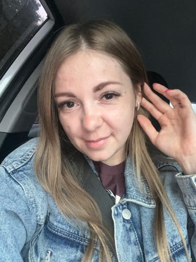 The image shows a close-up, selfie-style photograph of a young woman with long blonde hair, wearing a denim jacket over a burgundy shirt. She is in a car, as indicated by the seatbelt visible across her chest and the car window in the background. Her right hand is raised to her ear, and she is looking towards the camera with a slight smile. The lighting suggests it is daytime, possibly late afternoon or early evening. The background is dark and out of focus, with a hint of trees visible through the car window. There are no specific details that indicate the location in Allaki, Russia.