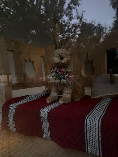 The image shows a straw rabbit decoration with a floral garland around its neck. The rabbit is positioned on a wooden structure that appears to be a small house or playhouse, possibly outdoors given the reflections of trees and sky. The rabbit is adorned with a pink and green ribbon and faux flowers. A red and white striped blanket or rug is draped over the side of the wooden structure. The overall scene evokes a rustic or autumnal theme. There are cutouts in the wooden structure, one shaped like a tulip. The lighting suggests it might be early morning or late afternoon, with soft light. The background shows a reflection of trees and a cloudy sky. No people or text are visible in the image.