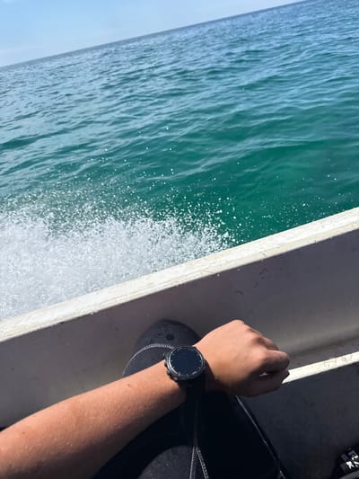 An arm wearing a black smartwatch rests on the side of a white boat. The boat is moving through clear, turquoise water, creating a spray of white foam. The sky above is a pale blue. The scene appears to be a daytime view from a boat on the sea, possibly near Limenaria, Greece, given the clarity of the water and the bright sunlight.