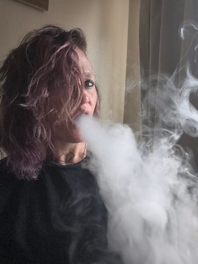 This image depicts an indoor scene in what is reported to be the city of Westminster, United States of America. A single adult individual is shown in a close-up, chest-up shot, viewed from a slightly right-of-center perspective. The subject has messy, medium-length hair with purple and pinkish hues, partially obscuring the right side of their face. The visible left eye features dark eyeliner and is directed forward. A substantial plume of dense, white vapor or smoke is being exhaled from the individual's mouth, extending diagonally across the lower right quadrant of the frame. A thin silver-colored chain necklace is visible around the neck, and a dark, angular tattoo is present on the left side of the neck, beneath the jawline. The individual is wearing a plain, dark-colored crew-neck garment. The background consists of a light-colored wall or curtain on the left, with a faint, dark rectangular object (possibly a framed item) on the upper left. To the right, a darker, semi-transparent curtain or window covering is visible.