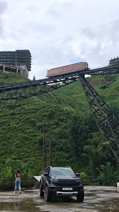 A low-angle shot shows a woman, a black Ford pickup truck, and a funicular railway in Cat Cat Village, Vietnam.
The woman, facing away from the camera, stands to the left on a wet, gray concrete surface near a low railing. She wears a cropped black top, blue jeans, and light-colored sneakers.
The black Ford truck is parked to the right of the woman. Its license plate reads "29K-179.83".
The funicular railway ascends a steep, green hillside in the background. A dark-colored metal framework supports the tracks. An orange and red funicular car is positioned on the tracks.
Behind the funicular, there is an unfinished concrete building and dense green foliage. The sky is overcast.