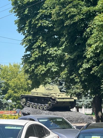 This image depicts a monument of a green tank, possibly a historical artifact, displayed outdoors in the city of Sarny, Ukraine. The tank is situated on a dark, rocky mound, which serves as its base. It is partially obscured by the dense foliage of a large tree with green leaves, suggesting a park or a public square setting. In the foreground, several cars are parked, with their roofs and windshields visible, indicating that the monument is located in an area accessible to vehicles and the public. The background shows more trees, a white fence, and a clear blue sky, consistent with an outdoor environment on a sunny day. There are no people visible in the image. The scene conveys a sense of stillness and memorialization, with the tank serving as a central point of interest within its natural and urban surroundings.
