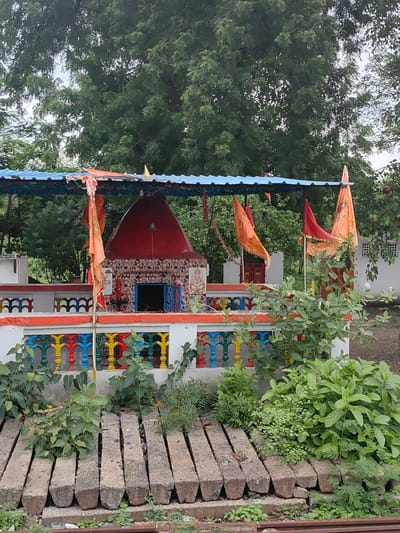 The image depicts a small, brightly colored Hindu temple located in Dujkheda, India. The temple features a red, conical roof and is decorated with floral tile work. In front of the temple is a white railing with colorful posts in red, yellow, green, and blue. Several orange and red flags are prominently displayed, attached to poles and draped over the temple's roof. To the left and right of the temple are white structures. The foreground is filled with lush green plants and several rectangular concrete blocks arranged horizontally. The background consists of dense green trees. The lighting suggests an overcast day. There are no people visible in the image.