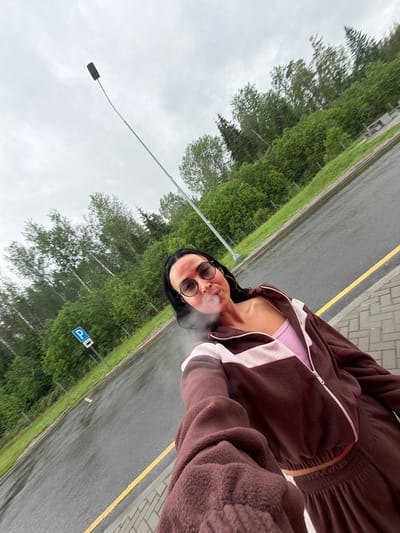 The image shows a person standing beside a street in Sust'e-Konets, Russia. The individual, wearing sunglasses and a brown fleece jacket over a pink top, is exhaling vapor, likely from an e-cigarette or similar device. Behind the person is a road with a yellow line marking, followed by a grassy area and trees. A street light and a parking sign are also visible. The weather appears overcast.