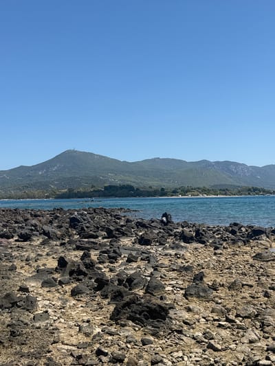 The image shows a rocky shoreline with a clear blue sky and a body of water. In the foreground, there are many dark, irregularly shaped rocks. The middle ground features the blue water with some ripples and a distant green shoreline with trees. Beyond the shoreline, a large, green, mountainous landscape is visible under the bright blue sky. There is a person in the water, swimming or wading. The location is the island of Lichada, Greece.