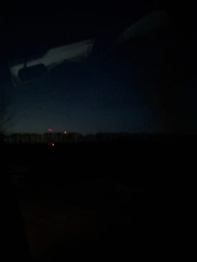 The image displays a nocturnal scene, observed from inside a vehicle, located in Primortsi, Bulgaria. The upper segment of the frame reveals the interior of the vehicle, featuring the silhouette of a rearview mirror suspended from the top center-left and blurred reflections on the windshield. The sky, visible in the upper mid-frame, is a deep blue, transitioning to black at the zenith. A dark, indistinct horizontal line, indicative of a distant tree line or terrain, traverses the mid-ground. Above this horizon, four distinct light sources are present: one prominent red light on the left, one prominent orange-yellow light further right, and two fainter, smaller orange-yellow lights situated below the brighter orange-yellow light and closer to the horizon. The foreground and lower mid-ground are obscured by darkness, providing no discernible environmental details. No individuals are visible, and no explicit actions or interactions are depicted.