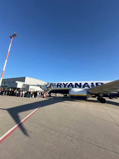 The image captures an outdoor scene at Weeze Airport, Germany, depicting a Ryanair airplane parked on the tarmac under a bright, clear blue sky. The aircraft's fuselage, emblazoned with the blue 'RYANAIR' text on a white background, dominates the upper right portion of the frame. A portable staircase is attached to the plane's door, leading to a visible entrance.
Below the plane, a large group of passengers is seen forming a queue on the tarmac, extending towards an airport building that runs parallel to the queue. The activity taking place is the boarding of the aircraft. Strong sunlight casts distinct shadows, including the outline of the airplane, onto the concrete ground which features red and white markings. This indicates a clear, sunny daytime. A red and white striped barrier pole is visible in the lower left corner against the vast blue sky.