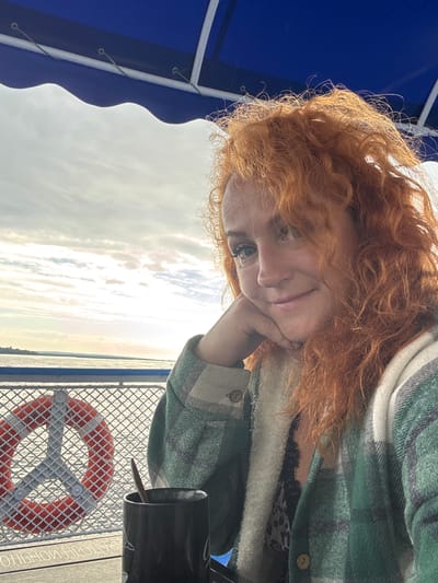 A woman with curly red hair takes a selfie on a boat. She is wearing a green plaid jacket over a black lacy top. Her right hand is resting on her chin as she smiles at the camera. In the background, a life preserver is visible on the railing of the boat, with the calm water of a river or lake behind it. The sky is overcast. The text "БОЛЬШОЙ ПРОГУЛОЧНЫЙ" which translates to "large excursion" is visible on the boat's railing, suggesting this is likely a pleasure boat tour. The location of Zuevo, Russia, cannot be determined from the image itself.