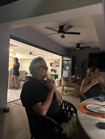 The image shows a patio with a view of the interior of a home. It is located in Playa Herradura, Costa Rica (latitude: 9.659, longitude: -84.672). The image is taken from the perspective of someone sitting at a table. Three people are visible in the scene: two are sitting at the table, while the third is standing in a room visible through a sliding glass door. The man seated to the left is wearing a black shirt and has his hands clasped together in front of his face. He is seated in a black wicker chair. The woman seated to the right is wearing a black dress and is looking at the man. She is seated in a black wicker chair. The woman standing in the room is wearing a black tank top and black shorts and is facing away from the camera. She is standing next to a counter. There is a ceiling fan in the room. The table is white with a black plate on it. There is an orange placemat on the table to the right of the plate. The table also has utensils and a cell phone. The floor is tiled and the patio has a sliding glass door that leads into the interior of the house. The interior is white with black accents. The ceiling fan and the lights in the room are visible. The patio has a black ceiling and white walls. The room beyond the sliding glass door has white walls. There are windows and a black chair in the room beyond the sliding glass door. There is a plant on the patio.