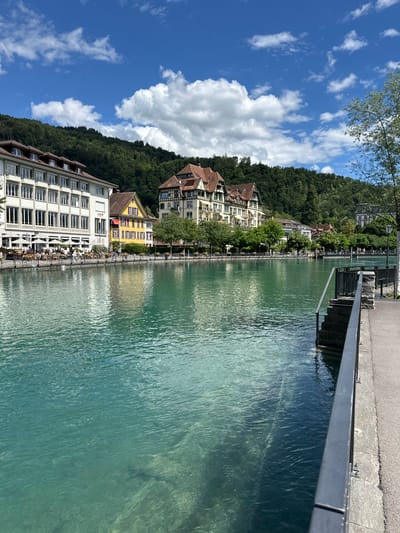 The image depicts a scenic view of the Aare River in Thun, Switzerland, on a clear day with scattered clouds. On the left bank of the river, a row of buildings lines the waterfront. The closest building is a large, multi-story establishment with a white facade and numerous windows, suggesting it is a hotel or a significant commercial building, indicated by the "AEK" logo visible on its side. Adjacent to it is a bright yellow building with red awnings. Further down the bank, other buildings with traditional Swiss architectural styles, featuring pitched roofs and decorative elements, can be seen. The riverfront is lined with trees and outdoor seating areas, implying a lively promenade or dining area.

The Aare River itself is a striking turquoise color, reflecting the bright blue sky and fluffy white clouds. The water appears clear, with some visible depth and texture from ripples on the surface. The right bank features a paved walkway with a dark metal railing. Stone steps lead down from the walkway into the river. A lamppost stands along the walkway. In the background, a densely forested hillside rises, characteristic of the surrounding landscape near Thun. Several people are visible strolling along the riverbank and seated at outdoor cafes, enjoying the pleasant weather and the picturesque environment. The overall impression is one of a tranquil and beautiful urban waterside setting.