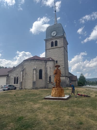 A full-shot image captures a scene in front of a stone church in Grande-Rivière Château, France. The church has a tall, square tower with a gray, shingled roof and a clock face on one side. A cross tops the tower. In the foreground, a large wooden sculpture depicts a man wearing a wide-brimmed hat, holding a stick, and standing next to a dog. The sculpture is placed on a wooden base. To the right of the sculpture, a man wearing a blue t-shirt and khaki shorts is walking past, with a red bicycle lying on its side in the grass nearby. The ground is covered in dry, yellowed grass. A silver car is parked to the left of the church. The sky is blue with scattered white clouds. In the background, a green hillside with trees is visible. Two other people are walking in the distance.