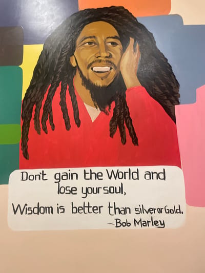 The image is a painting of Bob Marley, a Jamaican singer, songwriter, and musician, set against a colorful, abstract background. He is depicted from the chest up, wearing a red shirt. His iconic dreadlocks frame his face, and he is smiling with his mouth open. One hand is raised to his cheek. Below the portrait, a white speech bubble contains a quote attributed to him: "Don't gain the World and lose your soul, Wisdom is better than silver or Gold. - Bob Marley."
The exact location of this painting in Barakin Lerawi, Nigeria, is not specified within the image itself. Therefore, no further details about the specific environment or surroundings in Barakin Lerawi can be provided.