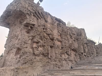 This is an upside-down, angled shot of the "Memory Cliff" memorial complex in Dubovka, Russia. The monument consists of a large, artificial rock formation with intricate carvings, inscriptions, and brickwork embedded within its structure. Steps are visible at the top left of the image. The sky is light and clear.
