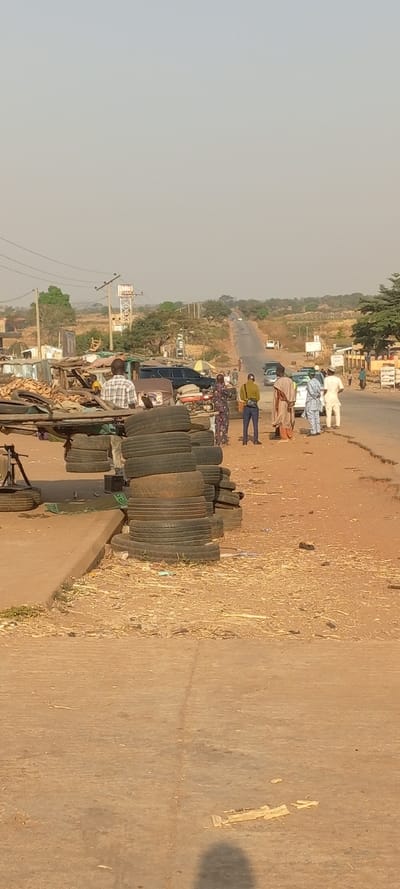 An outdoor scene shows people gathered near a road with vehicles in the distance. A stack of tires and a small market are in the foreground. The overall environment appears dry, with an earth-toned color scheme. A tower is visible on the horizon. The sky is hazy, suggesting the scene is taken during daylight hours.