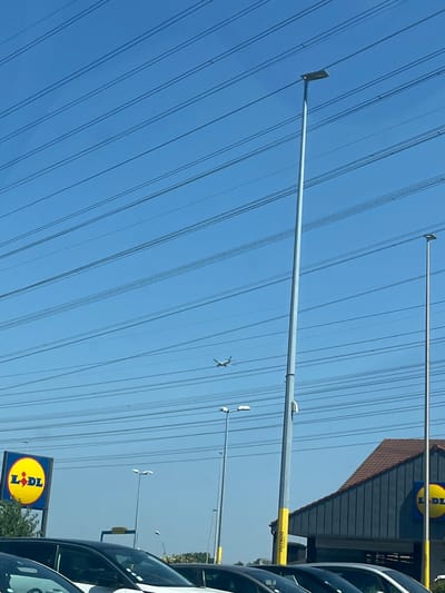 This is a daytime, eye-level, exterior shot taken in Villebon-sur-Yvette, France, under a clear blue sky filled with a complex network of overhead power lines. In the mid-ground, a commercial building with a dark blue facade and a brown tiled roof is visible. Two large, circular yellow signs with the "Lidl" logo are prominently displayed on the building's exterior. Several tall, slender grey lampposts with rectangular light fixtures stand throughout the scene, some with yellow accents at their bases. In the upper portion of the sky, a small airplane is in flight. In the foreground, the hoods and roofs of several parked cars are visible.
