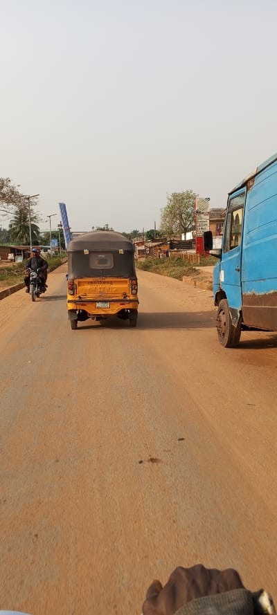 This image presents a street scene in Igbesa, Nigeria, viewed from a slightly elevated, first-person perspective, likely from a moving vehicle. The foreground and mid-ground are dominated by a wide, unpaved road with a reddish-brown, dusty surface, characteristic of laterite soil. The sky is a pale, hazy white, suggesting either an overcast day or a dusty, dry season atmosphere.
In the center of the road, a yellow tricycle, commonly known as a "Keke Napep," is moving away from the viewer. It features a black canvas canopy and is notably carrying several long, light-blue metallic structures, possibly corrugated roofing sheets or scaffolding, tied securely to its roof and extending upwards and backward. The rear of the tricycle clearly displays the manufacturer's name "PIAGGIO," along with "AYUWOPA" (likely a local transport association or operator name), and contact numbers "08133220116" and "07038166699". Its license plate reads "OSUN KSR-281-XC," indicating registration in Osun State, Nigeria.
To the right of the tricycle, the front section of a weathered blue truck or van is partially visible, also moving in the same direction. On the left side of the road, a person on a motorcycle, dressed in dark attire, is approaching the viewer.
The background and mid-ground reveal a semi-urban or rural landscape. Simple, single-story buildings, some appearing to be shops, line both sides of the road. Sparse vegetation, including trees and shrubs, is scattered throughout. Several utility poles or streetlights stand alongside the road. On the right, near the blue truck, a red and white signpost advertises various services, including "PMA MEDIX," "Coolmart," "Lubricants," and "Car Wash." A dark-skinned hand wearing a watch is visible in the bottom right corner, reinforcing the personal viewpoint of the photographer. The scene captures typical daily activity and transport in a Nigerian local setting.