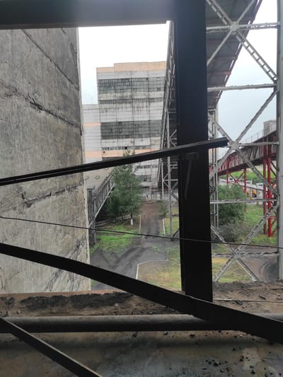 The image shows an industrial area, likely a former factory or processing plant, with a large concrete structure on the left and metal scaffolding and walkways in the foreground and background. A multi-story building with many windows, some broken, is visible in the distance. The ground is covered with dirt and debris, and there are some trees and vegetation. The sky is overcast. The scene suggests a derelict or abandoned industrial site. It is not possible to definitively identify this as being in Kyzyl, Kazakhstan, as there are no specific identifying markers or context provided within the image itself.