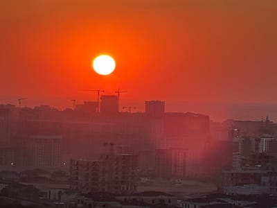 The image depicts a city skyline at sunset, bathed in a vibrant orange hue. The sun, a bright white orb, is prominently positioned in the upper center of the frame, casting a warm glow over the urban landscape. Numerous buildings, varying in height and architectural style, are visible, some appearing to be under construction, indicated by cranes and unfinished structures. The overall scene suggests a densely populated area, with a haze or dust creating a soft, diffused light effect. The location is identified as Kok Jar, Kyrgyzstan.