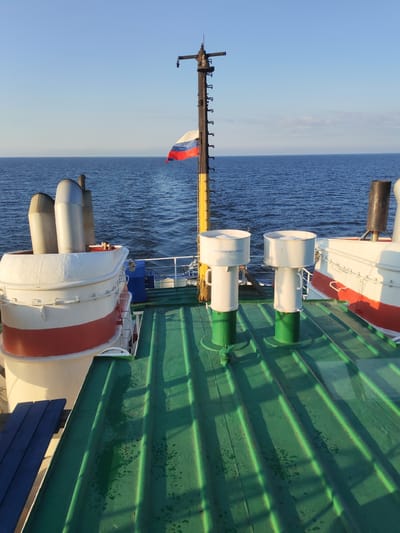This image depicts the deck of a vessel sailing on a body of water, likely the Volga River near Prislon', Russia, given the presence of the Russian flag. The perspective is from the deck looking towards the stern of the ship. In the foreground, a green-painted deck with raised ridges and some water droplets is visible. To the left, a white and red life raft structure with two large metallic exhaust pipes emerging from it dominates the scene. Further back, two white ventilation shafts with green bases stand on the deck. Behind these, a tall mast with a Russian flag attached to it is prominent. The background is filled with a calm blue body of water meeting a clear blue sky. The lighting suggests it is either early morning or late afternoon. No people are visible in the image.