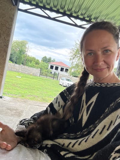 A woman is taking a selfie outdoors. She has light blue eyes and light brown hair pulled back in a braid. She is wearing a black and white patterned sweater. The background shows a grassy yard, a brick wall, a building under construction, a fence, and a white car. The sky is cloudy. It appears to be daytime. The woman's expression is neutral.