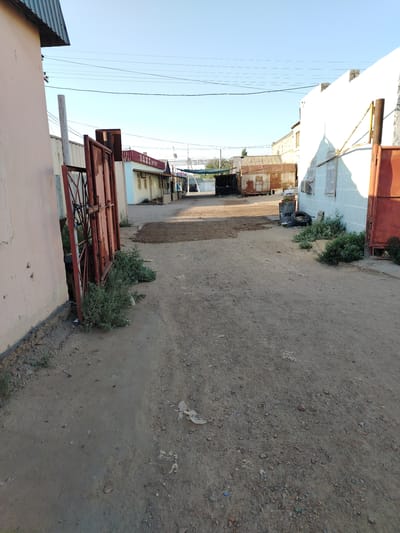 A dirt road lined with buildings and industrial elements leads into the distance under a clear blue sky. On the left, a pale pink building with a dark metal roof edge is visible, adjacent to a rusty red metal gate and overgrown green bushes. Further down the road, several structures with corrugated metal roofs and light-colored walls are present, some appearing to be shipping containers. A sign with Cyrillic lettering, possibly reading "DAMN AKKEN," is mounted on one of these buildings. To the right, a white building with blue trim features a large red metal door and an attached structure. Industrial debris, including tires and barrels, is scattered near the white building. The ground is covered with dirt, gravel, and some scattered trash. The scene appears to be an outdoor industrial or commercial area in the city of Töretam, Kazakhstan.