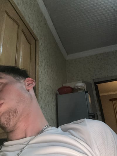 The image displays an interior room, viewed from a low angle looking upwards, featuring a male subject and various household items. The specific location within Durgena, Georgia, cannot be determined from the visual information provided.

**Subject:**
A male individual is partially visible on the left side of the frame, from the lower neck to the upper portion of his head. He has short, dark hair with a fade haircut on the left side and stubble on his cheek and jawline. A silver-colored chain necklace is worn around his neck, resting on a light-colored, likely white or light grey, t-shirt with a subtle textured pattern visible on the right shoulder area. His left ear is clearly visible, and his face is partially obscured, with his gaze directed downwards or away from the camera.

**Environment and Objects:**
The background consists of a wall covered in light-colored, possibly beige, patterned wallpaper. To the upper left of the subject, a light-brown wooden door with two arched panels and a distinct wooden trim is visible. The ceiling is light-colored, appears to be paneled or ribbed, and features white trim along the wall-ceiling junction.

To the right of the subject, a silver-colored appliance, consistent with a refrigerator or freezer, stands against the wall. On top of this appliance, a red plastic bowl or container is positioned on the left, next to a lighter-colored, possibly white or beige, lidded container or bowl on the right. Further to the right, a doorway reveals an adjacent room where a window with light-colored curtains is visible, suggesting an external light source or another room. The overall lighting is consistent with an indoor setting, showing shadows and highlights indicative of artificial illumination.