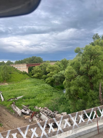 The image shows a rural landscape under a cloudy sky, likely in or near Новосёлки, Russia. In the foreground, a section of a wooden railing is visible, decorated with white tape forming an "X" pattern. Beyond the railing, to the left, are piles of concrete debris and exposed rebar, suggesting demolition or construction work. A dirt path runs alongside the debris. The middle ground is dominated by a lush green valley with a meandering stream. On the left side of the valley, a hill is covered in dense green vegetation. Spanning the valley is a bridge with concrete supports and a rusty red metal superstructure, appearing to be an older railway or road bridge. The background features a dense forest of green trees. The sky is overcast with dark gray clouds, hinting at impending rain. No people are visible in the scene.