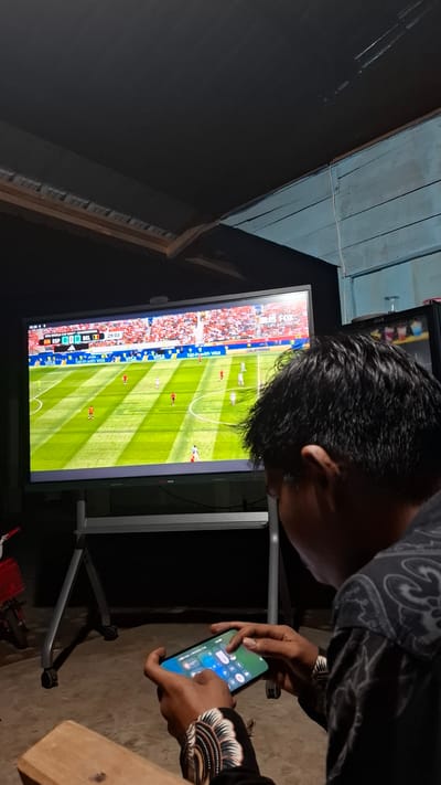 A person, viewed from behind, is holding a smartphone in their hands, interacting with the screen which displays a control panel with various icons. They are seated in a room with a large television screen in front of them, displaying a live soccer match. The room has a dark ceiling with wooden beams and a blue painted wall on the right. The floor is concrete, and a bicycle with a red basket is visible on the left side of the image. The scene appears to be in a residential setting in Grong-grong, Indonesia, likely a living room or a recreational space.