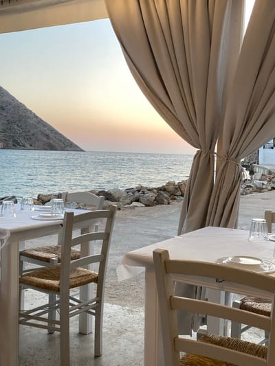The image depicts an unoccupied outdoor dining area situated directly on a paved coastal promenade during dusk. In the foreground, two white wooden dining tables are visible; the left table is fully shown, set with four white dinner plates and four inverted clear drinking glasses, accompanied by two white wooden chairs featuring woven straw seats. On the right, large beige fabric curtains are tied back, framing the view of the sea. Beyond the tables, a rocky shoreline borders the tranquil, blue-green sea. In the background, a large, treeless mountain slope rises on the left across the water. The sky displays a gradient of colors, transitioning from light blue overhead to warm orange and pink hues at the horizon, indicative of sunset. A portion of a whitewashed building is visible on the far right. The scene is located on the coast in Greece, near the city of Apollonia.