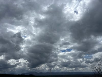 The image depicts a cloudy sky over a landscape. The sky is dominated by heavy, gray clouds with occasional patches of blue visible through openings. The landscape below features a rolling terrain of green fields and distant hills. There is a utility pole visible in the lower portion of the image, and some bare branches on the right side. The scene suggests a rural setting. There are no people visible in the image. The location is not explicitly stated in the image to be Srednia, Bulgaria, but is assumed based on external data.