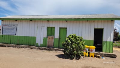 The image displays a single-story building under a bright blue sky with scattered white clouds. The building features a corrugated metal roof with a green trim and walls composed of vertical wooden planks, painted white on the upper section and green on the lower.

On the left side of the building, a white informational sign is prominently displayed. Logos for organizations such as "Solidarités International," "Solthis," and "ALIMA" are visible, alongside symbols representing the French government and the European Union. The sign's text, partially in French, indicates a project titled "Vision Provinciale de la Santé d'ITURI / Zone de Santé Rwampara," with further details like "Titre du Projet:", "O, SHARL MAKARO," and "RONDONLAMPSRA."

The building has multiple openings: a white wooden panel on the far left, a green wooden shuttered window or double door partly obscured by a wooden board, and a closed green wooden door next to it. To the right, an open doorway reveals a dark interior with a visible bench.

In the foreground, a large, dense green bush sits on the unpaved, reddish-brown dirt ground, casting a shadow. Next to the open doorway, two yellow lidded trash bins are visible, one clearly marked with the "ALIMA" logo. The overall scene suggests a functional, community-focused structure, likely in a rural or semi-rural setting in daytime. A "Joseph Kisuki" watermark is present in the bottom right corner of the image.