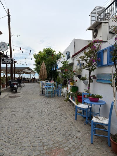 This image depicts a cobblestone street in Olympiada, Greece, lined with outdoor seating for a cafe or restaurant. On the left side of the street, a sign for "MARIANNI ROOMS" hangs above a satellite dish. A blue motorcycle is parked next to a table and chairs. Further down the street, there are more tables and chairs set up under a covered area, with people seated at some of them. The sea is visible in the background. On the right side of the street, a white building features blue shutters and potted plants adorning its exterior. Colorful string lights are strung across the street, adding a festive touch to the scene. The overall atmosphere appears to be one of a relaxed, seaside town.
