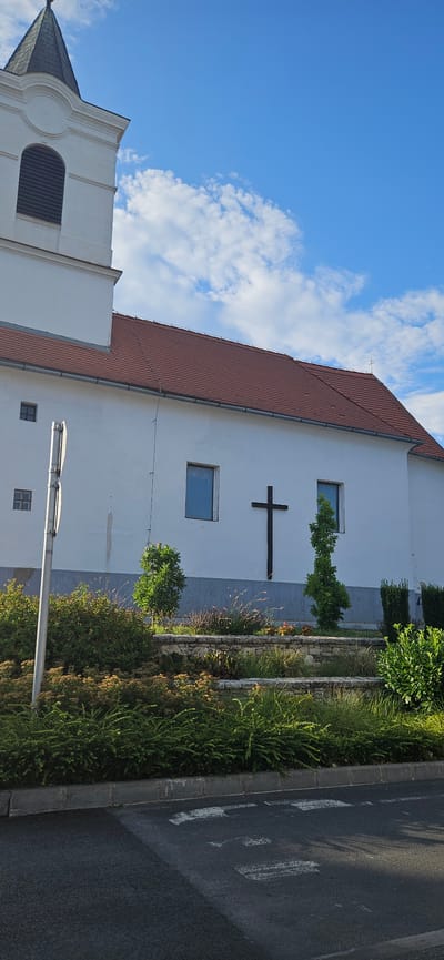 The image depicts the exterior of a white-walled church with a red-tiled roof in Balatongyörök, Hungary. A dark, wooden cross is prominently mounted on the church's facade between two rectangular windows. A white bell tower, partially visible on the left, extends upwards and is topped with a dark spire. The building is situated in front of a bright blue sky dotted with scattered white clouds. In the foreground, a landscaped area features various types of greenery, including bushes and small trees, arranged on tiered stone walls. A grey paved road with white markings is visible at the very bottom of the image. The scene is illuminated by natural daylight.
