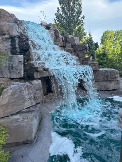 This media file captures a vibrant waterfall cascading over a substantial rock formation under a bright, clear sky in Chelsea, Canada. The primary subject is the waterfall itself, where light blue-green water vigorously tumbles down multiple tiers, creating abundant white foam and spray, particularly in its upper and middle sections. The water appears turbulent as it flows, settling into a calmer, but still foamy, pool at the bottom.

The waterfall's structure is composed of large, angular rocks, ranging in color from dark grey to brownish-grey. Many of these rocks exhibit a blocky or columnar appearance, forming distinct ledges and steps for the water's descent. Some sections of the rock face are dark and wet, while others show drier textures. Small patches of green moss or vegetation cling to crevices and edges of the rocks.

Above and around the rock formation, a variety of green trees are visible, including both dense evergreen conifers and lighter-foliaged deciduous trees, indicating a natural or well-maintained park-like setting. The bright, clear sky suggests it is daytime with sunny weather. There are no people or discernible text visible in the image. The scene evokes a sense of natural beauty, dynamic motion, and tranquility.