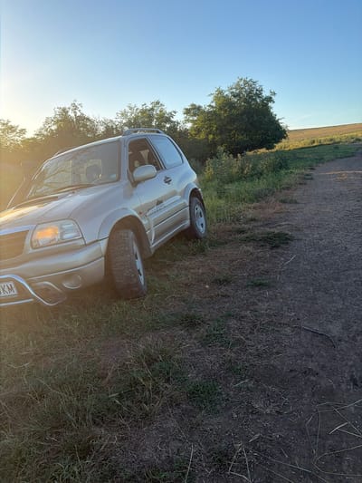 An outdoor, eye-level, medium shot captures a tan 2003 Suzuki Grand Vitara parked on the side of a dirt road. The vehicle is facing towards the left of the frame and is positioned on an upward slope. The car's headlights are on, and its front bumper features a chrome grille guard. The car's front license plate is visible, displaying "6989 KM". The surrounding environment is a rural area with dry grass, green vegetation, and trees. In the background, a cultivated field stretches towards the horizon under a clear blue sky, illuminated by the warm glow of the setting sun. The photograph was taken in Kozlovets, Bulgaria.