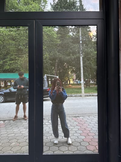 The image shows a reflection in a glass door, likely of a storefront. The reflections depict two individuals.
The top reflection shows a person wearing jeans, a blue jacket, and sneakers. They are holding a phone up to take a picture. The background behind them includes trees, a road with patterned paving stones, and utility poles.
The bottom reflection shows a person wearing shorts and a dark hooded sweatshirt, also using a phone. In the background, a car is visible, along with a storefront awning with some text.
The scene is likely Pejë, Kosovo due to information in the other images.