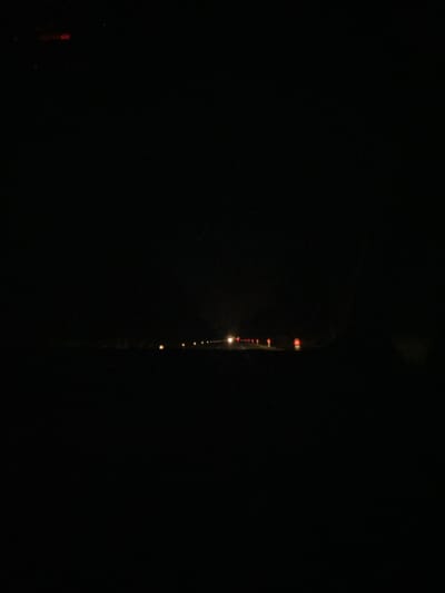 The image depicts a dark, rural road at night. A faint line of lights, likely from distant houses or streetlights, stretches across the horizon. In the foreground, there are some illuminated markers or traffic cones. The overall impression is one of deep darkness, with only scattered points of light breaking the gloom. The image does not provide enough detail to determine the specific location within Horbów-Kolonia, Poland, or to identify any people, subjects, or specific actions.
