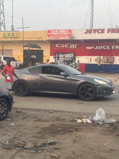 A gray Hyundai Genesis Coupe sits on a dirt road. A man in a red shirt stands near the car. Trash litters the roadside. In the background, a building is adorned with advertisements for "JOYCE FR" and "C&C INC. BRANCH #1," which distributes 6S PLUS BREWERIES INC. There's also a "BIG DADDY" visual/energy mix advertisement. The overall setting is a commercial street in an urban area.