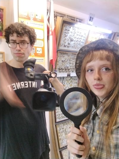 A person with dark curly hair and glasses holds a video camera. The person wears a black shirt with the word "INTERNET" printed on it. Standing next to them is a young woman with blonde hair and bangs, wearing a bucket hat and plaid shirt. She holds a magnifying glass. Behind them is a wall filled with framed displays of butterflies. A poster and a flag are also visible in the background. The woman is sticking her tongue out slightly.