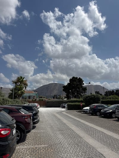 The image depicts a street scene in Carini, Italy, with a row of parked cars lining a cobblestone street. The cars are various models and colors, including black, dark red, and white. The street is bordered by a manicured hedge and trees. In the background, a large, rocky mountain rises against a bright blue sky filled with fluffy white clouds. A few buildings with orange roofs are visible near the base of the mountain, suggesting a residential or commercial area. One building appears to be under construction with scaffolding. A palm tree is also visible in the mid-ground. The time of day appears to be daytime, with bright sunlight casting shadows. The weather is clear and sunny, with a few dramatic clouds. There is no visible text in the image. The scene suggests a quiet, possibly Mediterranean, setting.