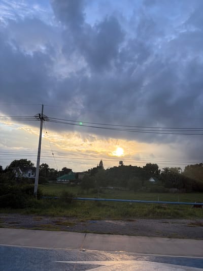The image depicts a serene landscape in Ban Ton Kwen, Thailand, during what appears to be either sunrise or sunset. The sky is dominated by dramatic, dark clouds that are beginning to break, allowing the golden rays of the sun to stream through, creating a beautiful contrast.

In the foreground, a paved road or pathway with a distinctive blue surface is visible, reflecting the ambient light. Adjacent to this is a gravel area, and further back, a grassy field stretches towards a line of trees and vegetation.

A prominent utility pole with electrical wires stretches across the frame, bisecting the sky and the landscape. The wires are supported by insulators attached to the pole.

Beyond the field, several structures are discernible. A white house with a distinctive roofline is situated to the left, partially obscured by trees. To the right of this, a building with a green, angled roof, possibly a mosque or a temple, stands out. Further in the distance, amidst the dense greenery, more homes and tropical vegetation are visible. A tall, slender tower, likely a water tower, is positioned atop a hill in the center-right of the image. A blue pipe runs horizontally across the middle ground, possibly part of an irrigation or water system.

The overall impression is one of rural tranquility, with the dramatic sky adding a touch of awe to the natural beauty of the scene. There are no people or animals visible in the image, and no specific actions or interactions are depicted.