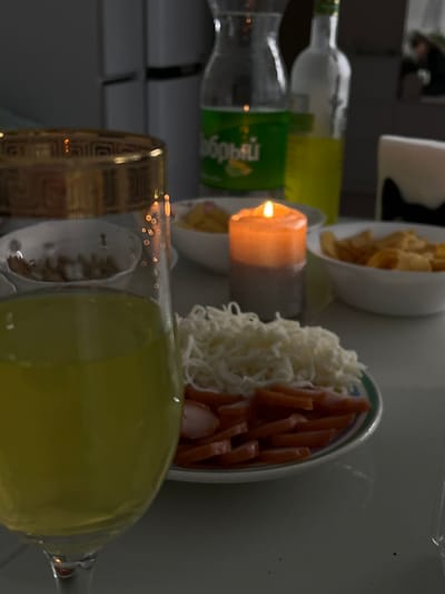 A candle is lit next to plates of shredded cheese, sausage slices, and chips. Bottles of clear liquid are behind the candle. In front of the candle, a fancy glass filled with yellow liquid stands on its side.