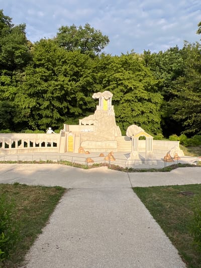 The image shows a miniature architectural model resembling classical ruins set amidst a park. The model is constructed from light-colored material and features structures with columns, arches, and steps. Two small, white human figures are visible on the model, one standing atop a wall and another lying down. The model is surrounded by a paved walkway and grassy areas. In the background, a dense line of green trees creates a natural backdrop. The sky above is partly cloudy, with soft, diffused light suggesting it is either early morning or late afternoon. The specific location in the city of Fontaines, France, is not evident from the image itself.