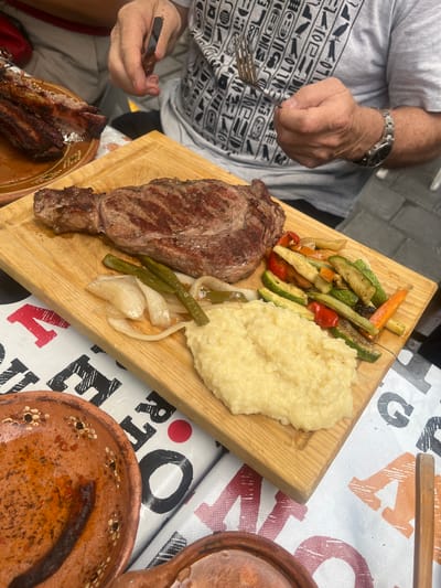 A man is seated at a table with a meal of steak, mashed potatoes, and grilled vegetables served on a wooden board. The steak is a large cut, and the vegetables include zucchini, bell peppers, and carrots. The table is covered with a patterned tablecloth. The man has a fork in one hand and a knife in the other. San Francisco Totimehuacán, Mexico, is not discernable in the image.