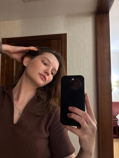 This is a portrait taken in a selfie format, featuring a young woman with long brown hair and fair skin. She is wearing a brown, textured sweater. A silver necklace with a small cross pendant is visible around her neck. Her left hand is raised, holding a black smartphone to capture the image, while her right hand is resting near her head. She appears to be indoors, with a wooden-framed mirror or wall visible in the background. The lighting suggests it could be daytime, but no specific time is discernible. The overall mood is casual and relaxed. There is no visible text in the image.