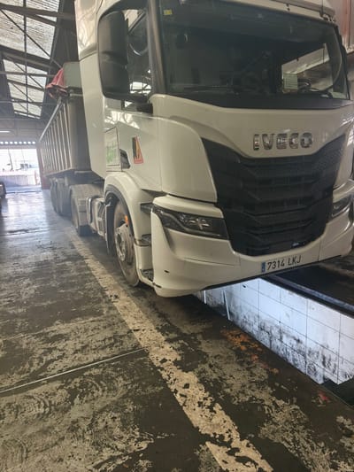 A white Iveco truck is positioned over a service pit in what appears to be a garage or workshop. The truck has a trailer attached and is facing the camera. The license plate "7314 LKJ" is visible on the front. The location appears to be indoors, as indicated by the overhead structure with metal beams and translucent roofing panels. The floor is marked with painted white lines, and there are stains and signs of wear and tear. The pit itself has tiled walls. The truck's tires are dirty and show signs of use. Based on the license plate and contextual clues, the location is likely in Spain. Barri De Porta, Spain, is plausible given this context, as well as the license plate format.