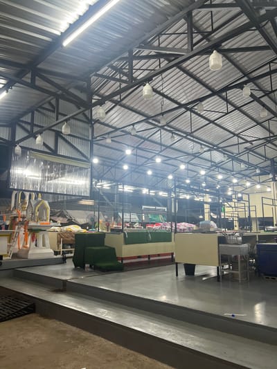 This image depicts the interior of a large, covered market structure, likely a food or goods market, in Thung Tako, Thailand. The scene is brightly lit by numerous overhead lights, some of which are fluorescent tubes and others appear to be dome-shaped lamps. The roof is constructed of corrugated metal, supported by a dark metal framework.
In the foreground, there is a grey concrete floor with tiered levels and drainage grates. To the left, a small, ornate altar or shrine is visible, adorned with garlands of orange flowers and various decorative elements. Behind the altar, a table displays a collection of items, including what appear to be textiles and some decorative pieces.
Further into the market, there are stalls or stands with counters. One counter is covered in a green artificial grass material, topped with more goods. Another counter is a light beige color, with a stainless steel prep area and cooking pot. Several fans are visible, suggesting a need for air circulation. In the background, rows of metal shelving units are present, holding various items, some covered in fabric. The overall impression is one of a bustling marketplace, likely operating in the evening or at night due to the prominent artificial lighting.