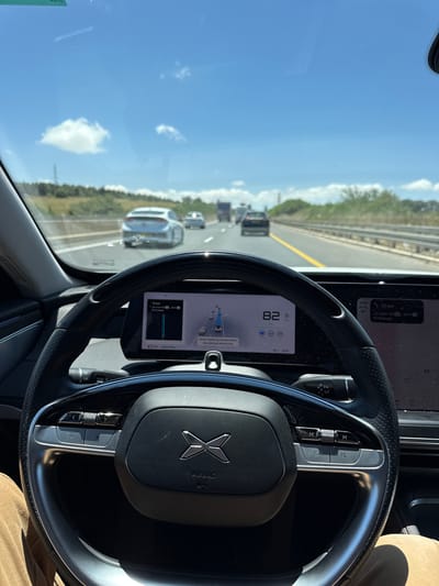 The image shows the interior of a car, specifically the driver's perspective. The steering wheel dominates the foreground, with the Polestar logo clearly visible. Behind the steering wheel, the car's dashboard display shows a navigation map, speed of 82, time of 12:54, and other vehicle information. Through the windshield, a highway is visible with multiple cars in motion. The sky is blue with scattered clouds. According to the dashboard, the navigation system is set to a destination 13km away in יקנעם עילית, which is located in the Jezreel Valley Regional Council, Israel. The driver monitoring system appears to be active.
