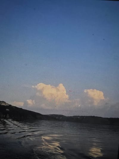 This image depicts a serene landscape with a body of water in the foreground and hills or cliffs in the background. The sky above is a gradient of blue, with some soft, yellowish clouds. The water appears to be calm with gentle ripples reflecting the light from the sky and clouds. The hilly terrain on the horizon is dark, suggesting it's either shaded or covered in vegetation. The scene captures a tranquil natural setting. This image is not taken in Dubno, Ukraine.