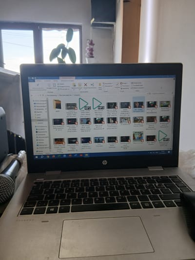 Here is a description of the image:
**Scene:** A close-up shot of an open laptop on a surface, likely indoors.
**Objects:**
* **Laptop:** An HP ProBook, keyboard visible with some wear, showing a Windows file explorer screen filled with various audio and video files. An external mouse is connected to the laptop.
* **Microphone:** A microphone is visible in the lower part of the image.
* **File Explorer:** The screen displays files related to songs and music videos.
* **Window:** A plant is visible in the background behind a window.
**Environment:**
* Appears to be an indoor setting with a window providing natural light, possibly a home or office.
**People:**
* No people are visible.
**Location:**
* The laptop's screen contains Cyrillic text, and the files on the screen suggest a relation to music. Text on the screen indicates "Младежко" (Mladezhko), which could be related to the location or theme of the files. The text on the screen indicates that the laptop is set to Bulgarian.